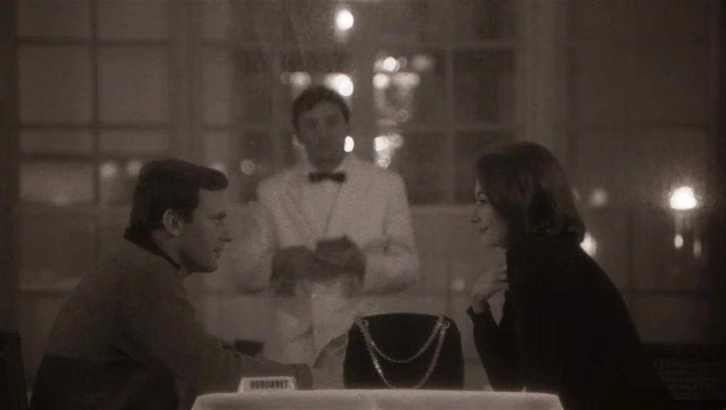 Un Homme et Une Femme (1966) movie still: medium — A dimly lit restaurant interior features a table with a small signage plate. A man and woman sit…