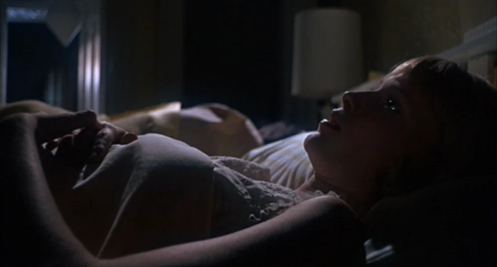 Rosemary’s Baby (1968) movie still: closeup — A dimly lit bedroom at night with a bedside lamp casting long shadows. A young woman lies in bed,…