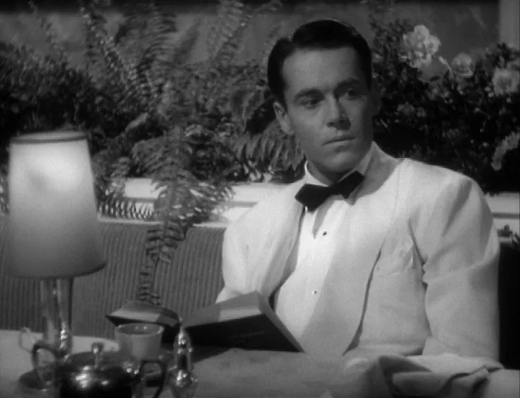 The Lady Eve (1941) movie still: medium — A man sits in a booth at a restaurant featuring potted ferns behind him. He holds an open book while…