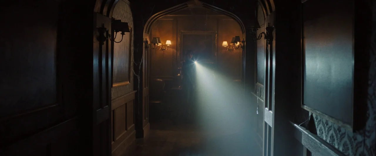 Abigail (2024) movie still: wide — A dimly lit gothic mansion corridor with wood paneling and aged wallpaper. A person stands in the distance…