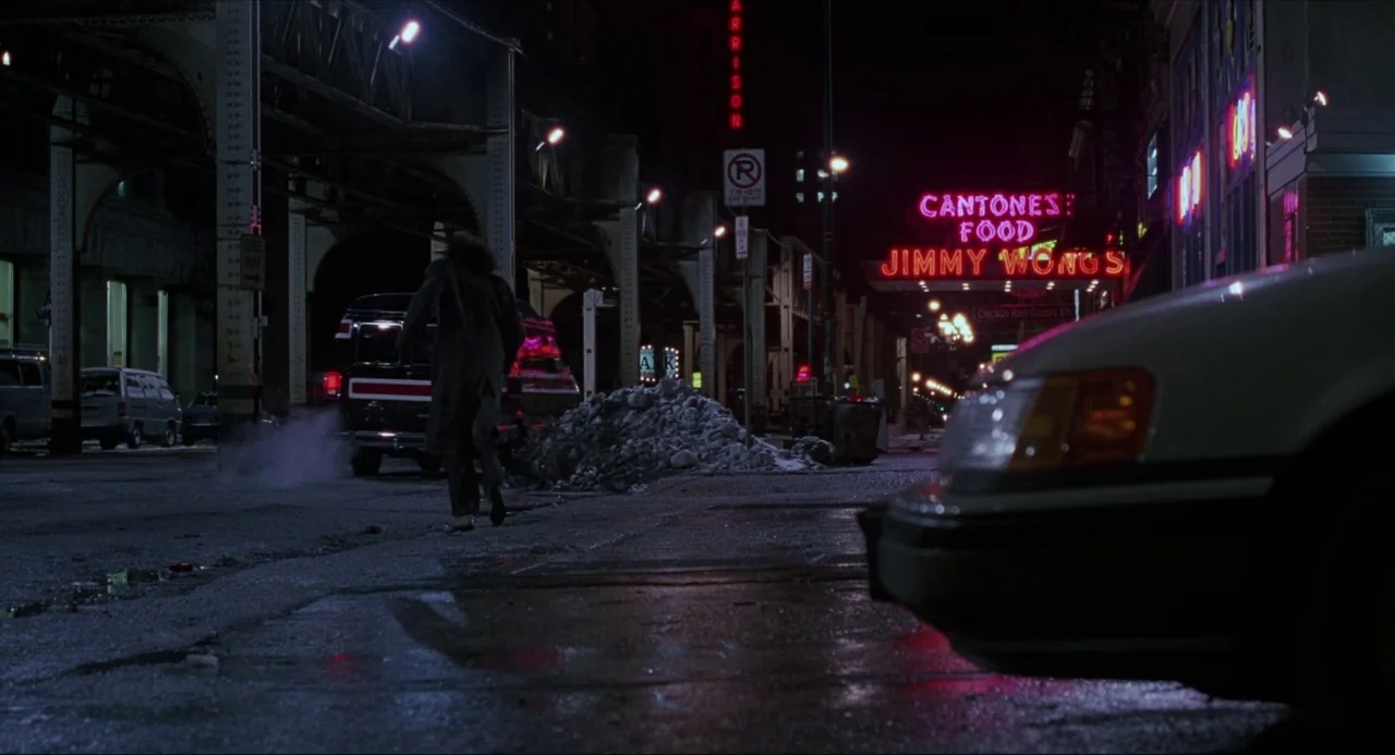 Child’s Play (1988) movie still: wide — An urban night street with overhead train tracks and multiple bright glowing neon signs. A man in a dark…