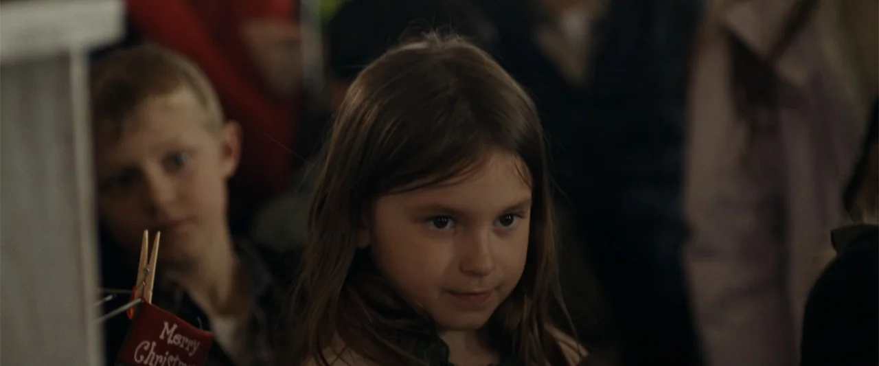 Silent Night, Deadly Night (2025) movie still: closeup — An indoor domestic setting features a young girl and a blurred boy behind her. The girl…
