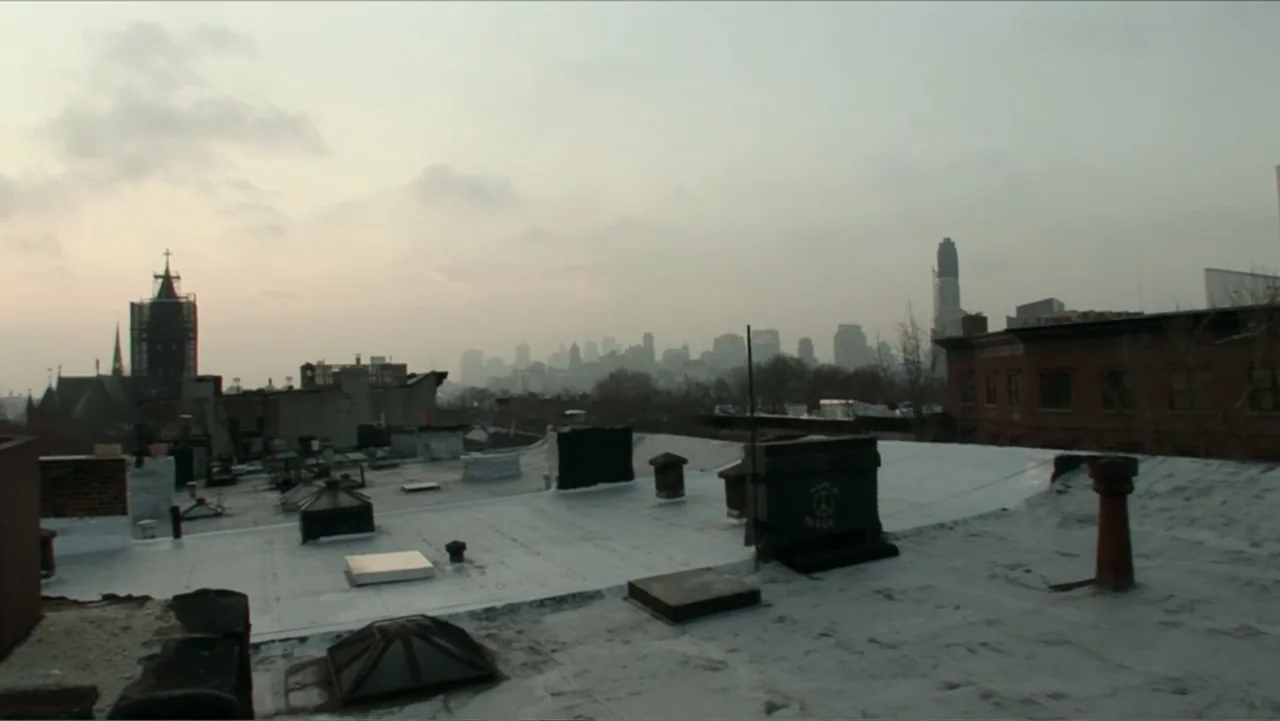 Little White Lie (2008) movie still: establishing — A flat tar-covered urban rooftop overlooks a hazy city skyline under an overcast sky. Several…