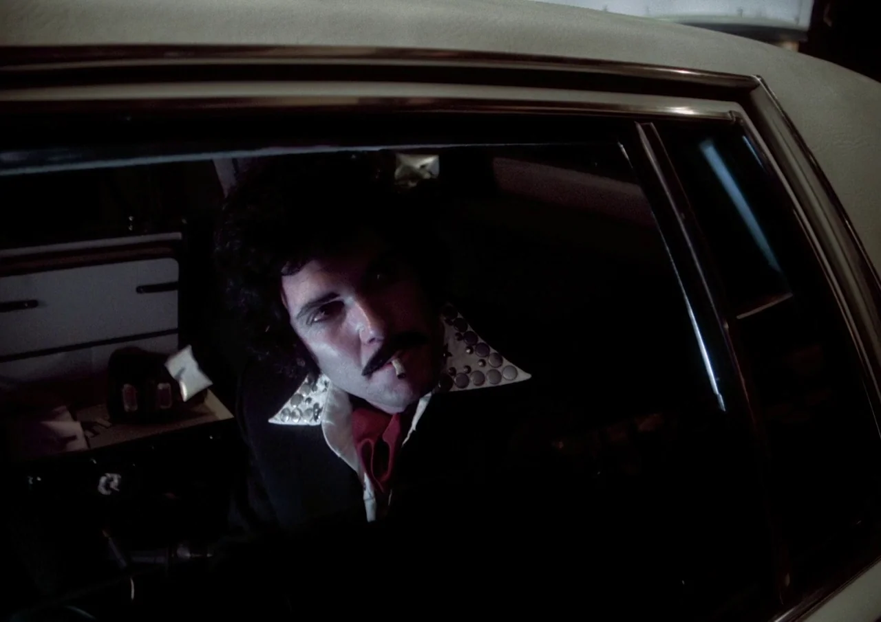 Human Highway (1982) movie still: closeup — A dark vehicle interior with the man framed through the open car window. The man sits in the driver seat…