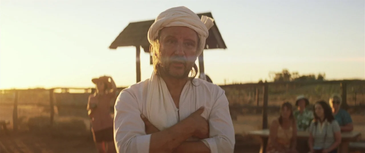 Tracks (2013) movie still: medium — A man wearing a turban stands outdoors in a dry, sunlit rural field. Several figures stand in the blurred…