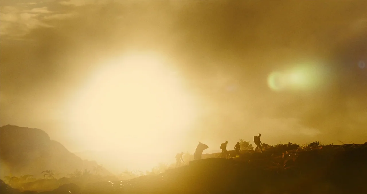The Suicide Squad (2021) movie still: wide — A group of people climbs a grassy, uneven ridge at sunrise or sunset. The intense golden sunlight…