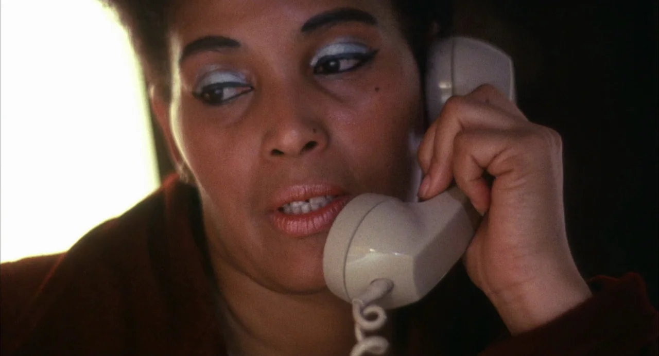The Killing of a Chinese Bookie (1976) movie still: closeup — A woman stands in a dim room with light filtering from a side window. She speaks while…