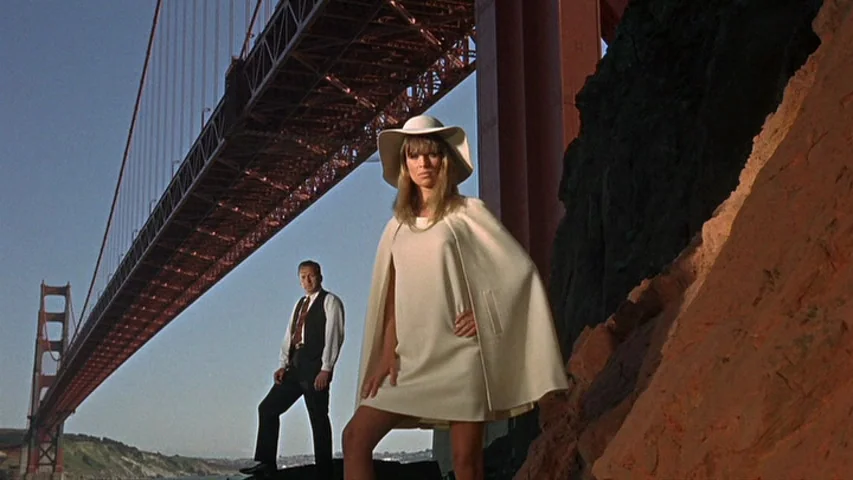 Petulia (1968) movie still: fullbody — A rocky shoreline sits directly beneath the steel frame of the Golden Gate Bridge. A woman in a white cape…
