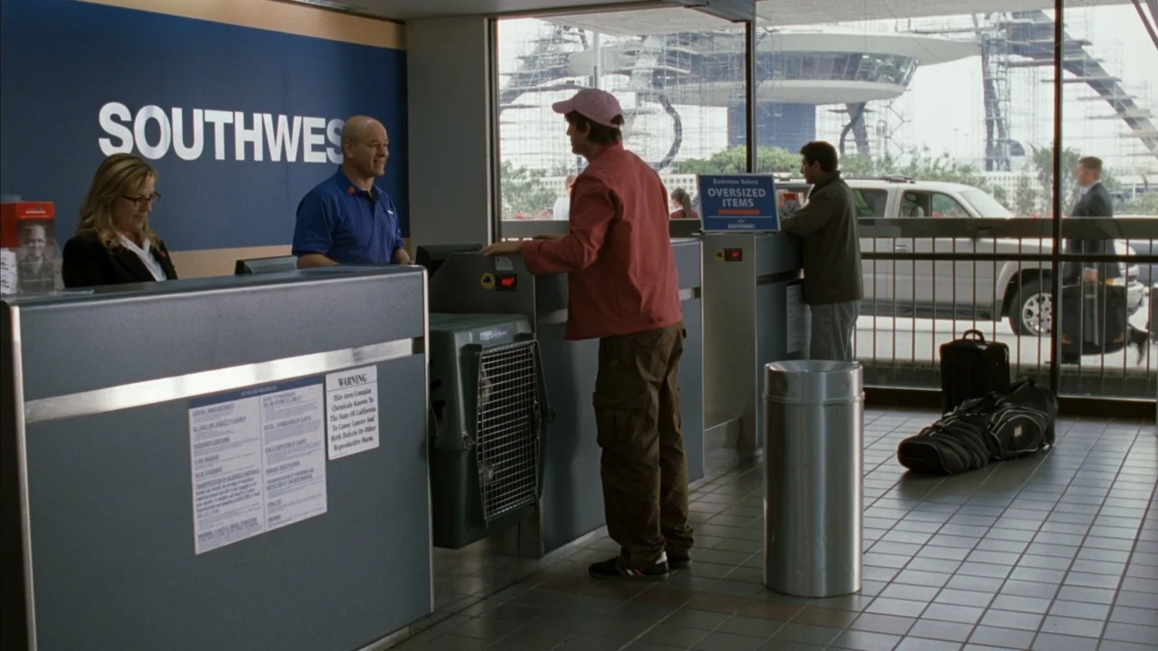 F Valentine's Day (2026) movie still: medium — An airline check-in area features a large blue counter with 'SOUTHWEST' branding in white text. A…