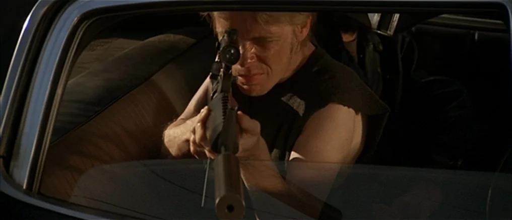Assault on Precinct 13 (1976) movie still: medium — A man sits inside a dark vehicle aiming a rifle at a target. He gazes down the sight of the…