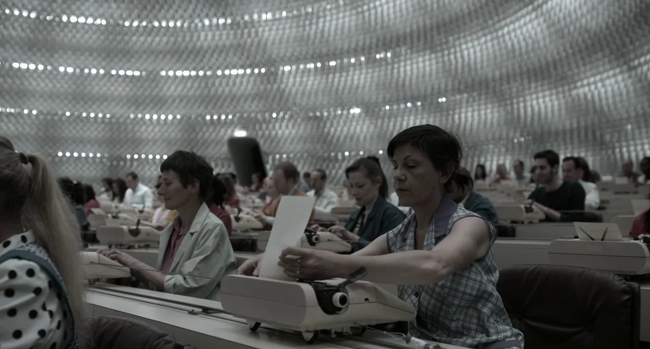 Mood Indigo (2013) movie still: medium — A large, semi-circular room with a textured, metallic ceiling and long rows of desks. Several individuals…