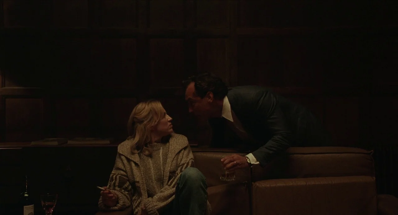The Nest (2020) movie still: medium — A dimly lit room features dark wood paneling covering the walls. A man leans over the back of a sofa toward a…