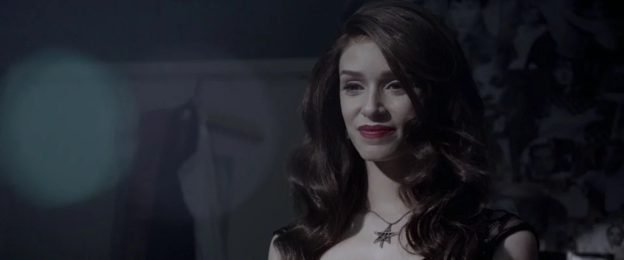 Starry Eyes (2014) movie still: closeup — A dimly lit indoor space features a wall with collage-style photographic prints. A young woman with wavy…