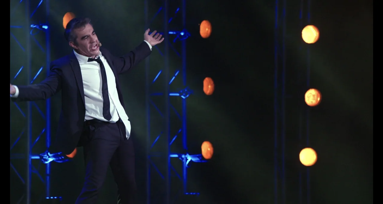 Paired Up (2019) movie still: fullbody — A man stands on a darkened stage illuminated by vertical rows of orange lights. He wears a formal suit and…