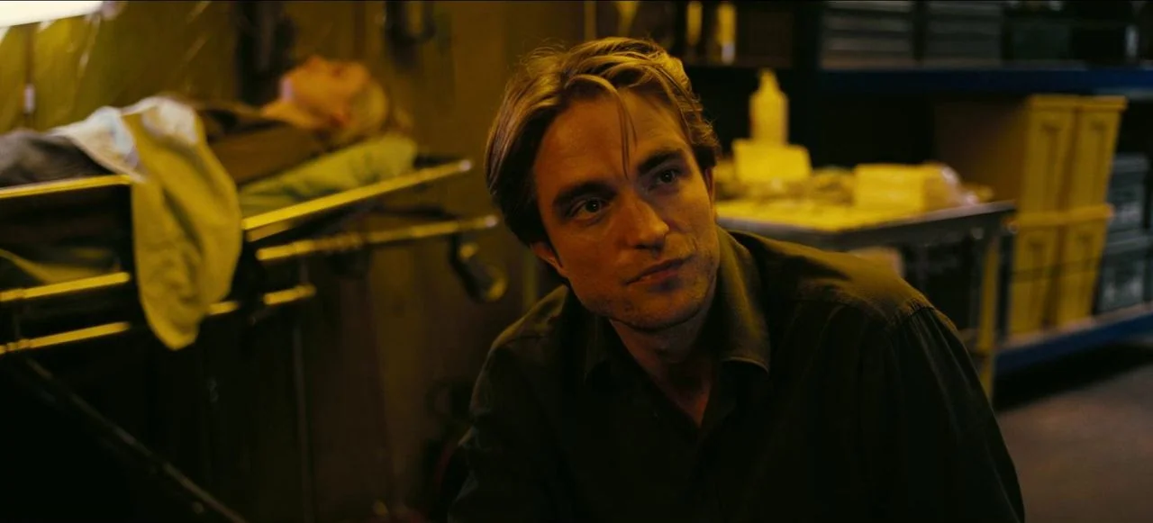 Tenet (2020) movie still: medium — A dimly lit storage space contains medical equipment and boxes on metal shelving units. A man sits in the…
