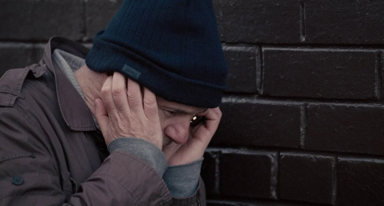 I, Daniel Blake (2016) movie still: closeup — A man stands against a dark, textured brick wall in an urban setting. He grips a small mobile phone…