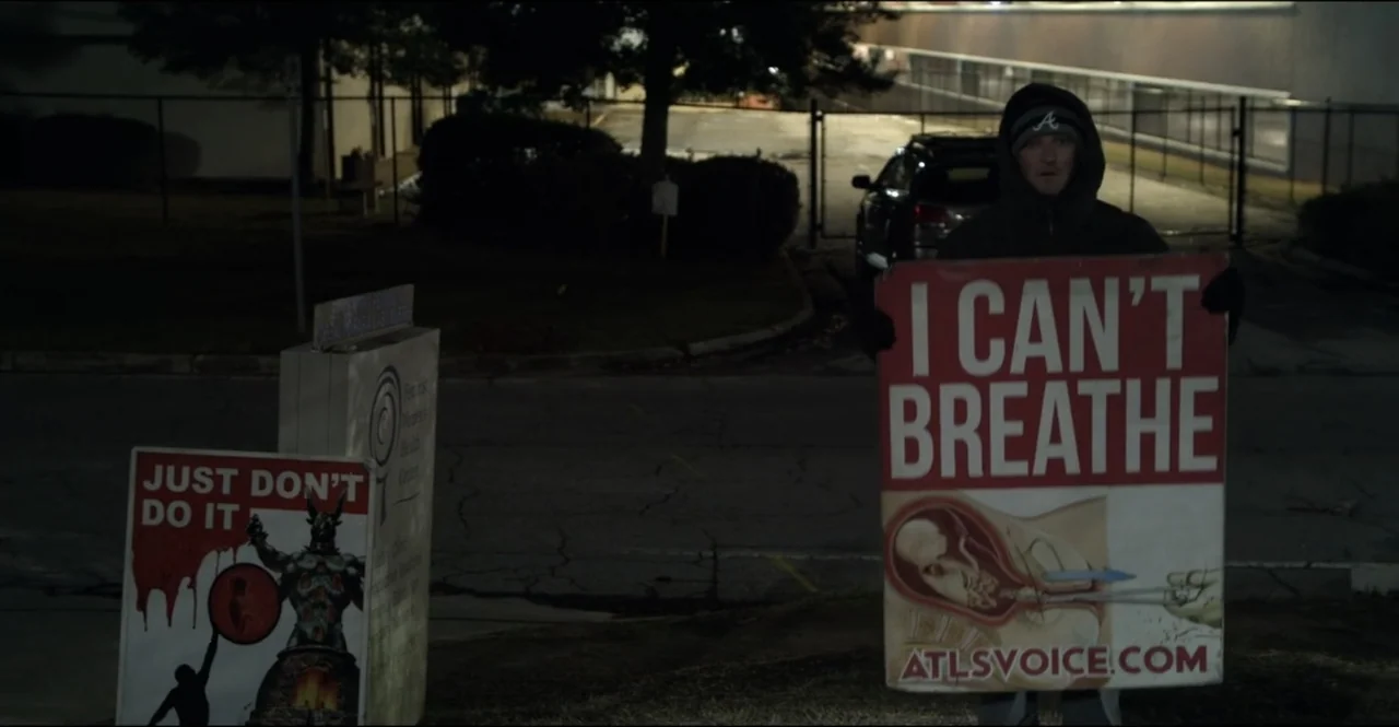 The Devil Is Busy (2024) movie still: medium — A man stands in a dark parking lot at night holding a protest sign. He wears a dark hooded jacket and…