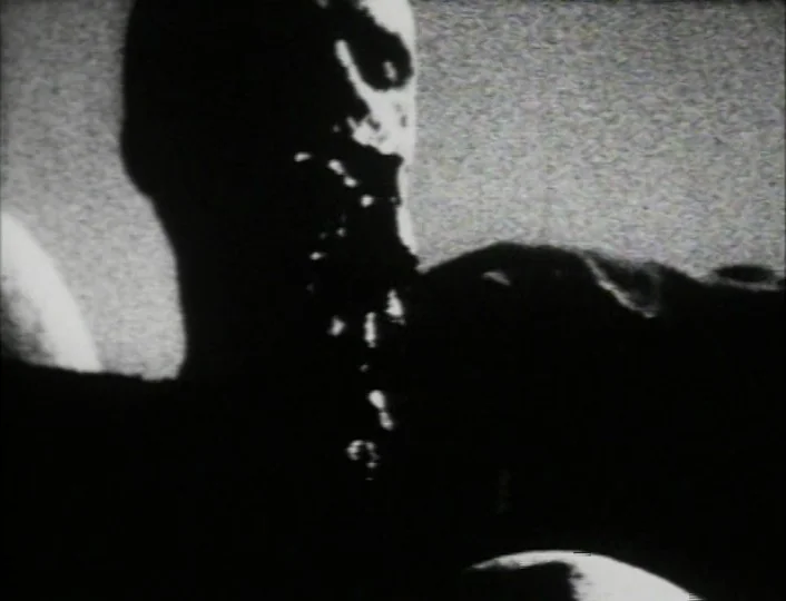 Begotten (1989) movie still: closeup — A low-resolution, high-noise frame depicting a shadowy figure in an unknown, dark space. The figure is…