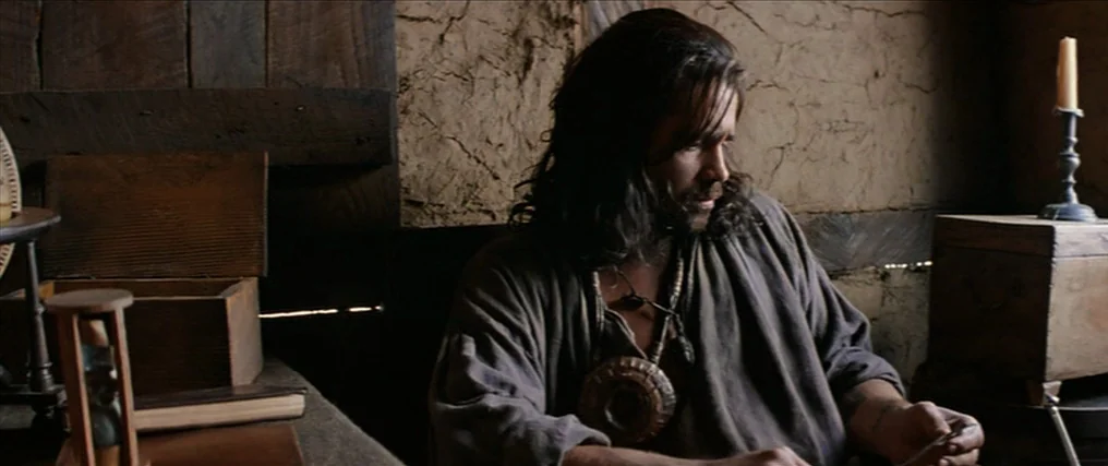 The New World (2005) movie still: medium — A dark wooden room with mud-plaster walls contains primitive furniture and a single candle. A bearded man…