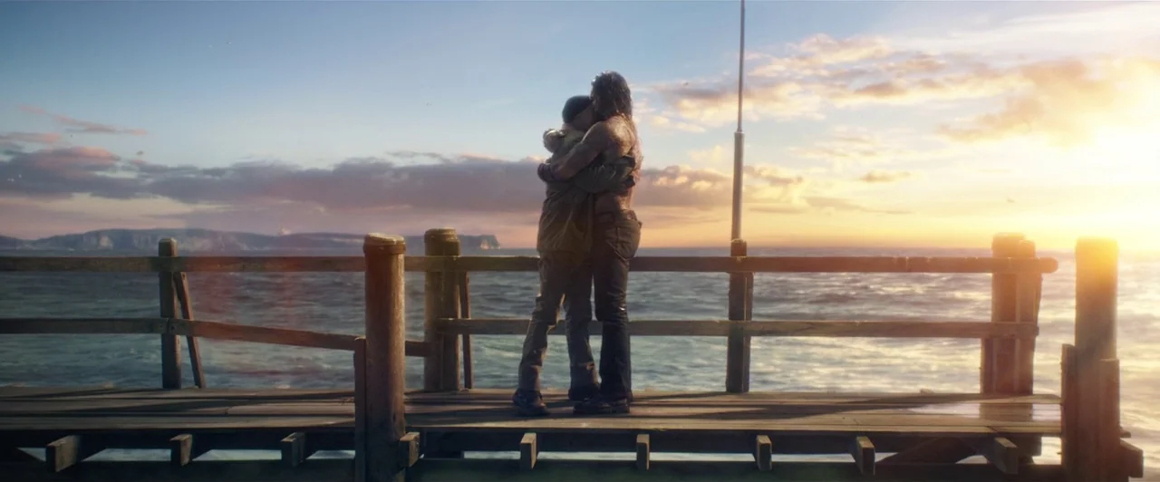 Aquaman (2018) movie still: fullbody — A wooden pier extends into a calm ocean during a bright golden sunset. Two figures stand together, embracing…