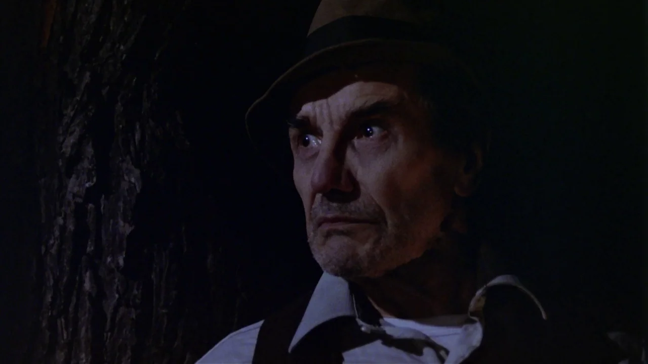 Friday the 13th Part 2 (1981) movie still: closeup — A man stands near a large tree trunk in a dark outdoor setting. He looks toward the side of the…