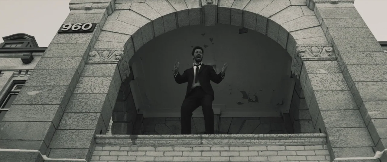 The Editor (2014) movie still: fullbody — A stone building entrance with an archway features the number 960 on its face. A man in a suit stands…