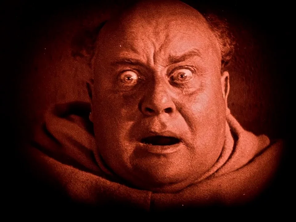 Häxan (1922) movie still: closeup — A dark, vignetted frame features a close-up of a bald man's face. The man expresses surprise with wide eyes and…