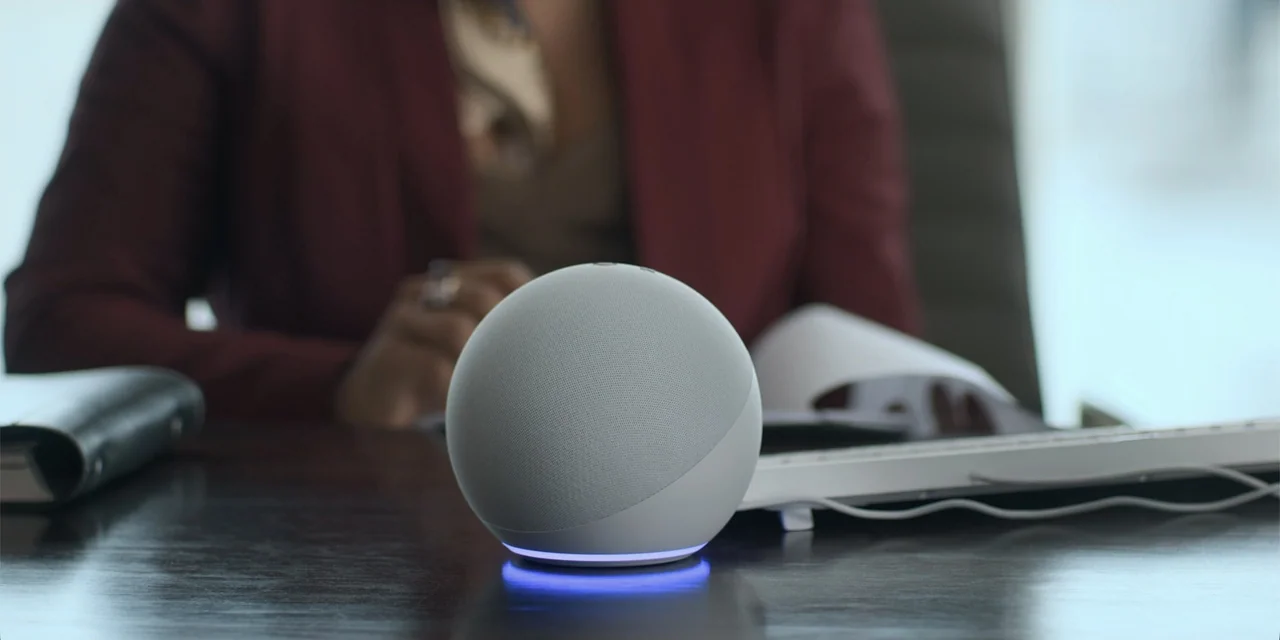 An Unforgettable Year - Autumn (2023) movie still: closeup — A modern office desk with a grey fabric smart speaker in the foreground. A person…