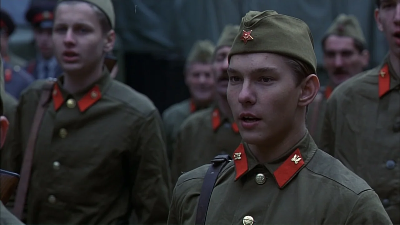Citizen X (1995) movie still: medium — A group of soldiers stand in a military encampment outdoors. A young man in a uniform speaks while the others…