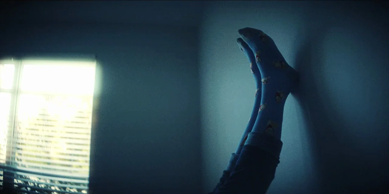 Spermworld (2024) movie still: medium — A dimly lit room features a window with blinds casting a strong light source. A pair of legs wearing…