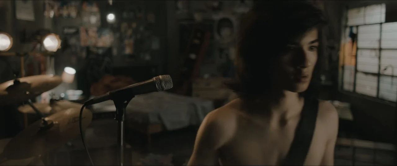 Death to Summer (2021) movie still: medium — A dark bedroom cluttered with musical instruments and posters on the walls. A shirtless teenager stands…