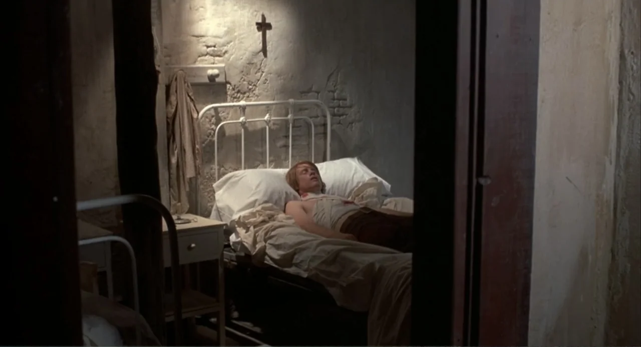 The Spikes Gang (1974) movie still: medium — A spartan room features a white metal bed against a rough textured wall. A man lies in the bed with his…