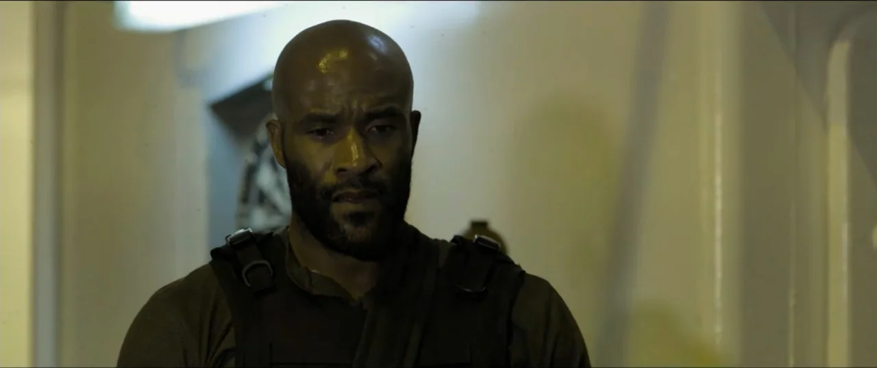 Primal (1994) movie still: medium — A dimly lit industrial corridor with neutral-toned walls and tactical equipment visible on walls. A man in a…