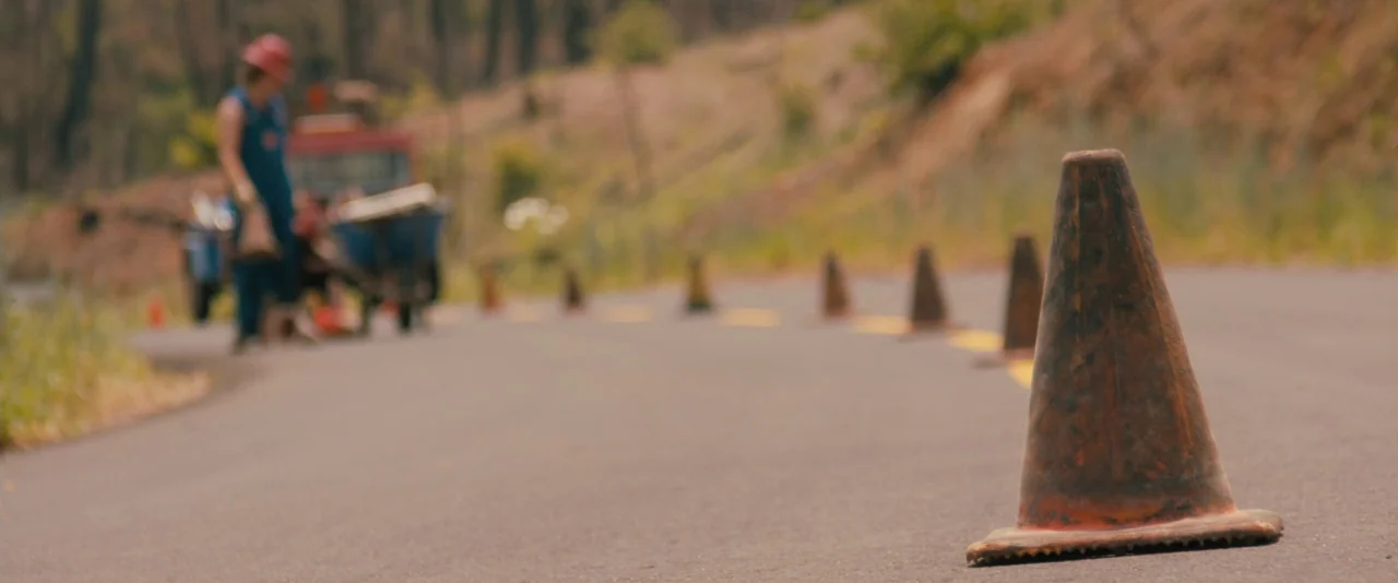 Prince Avalanche (2013) movie still: wide — An asphalt road winds through a rural landscape with a dirt shoulder. A singular orange traffic cone…