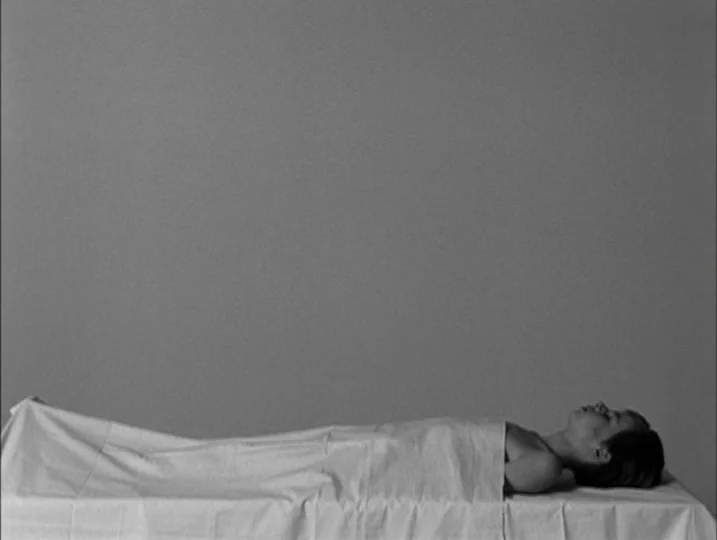 Persona (1966) movie still: wide — A minimalist, empty room with a plain grey wall behind a white examination table. A person lies supine on the…