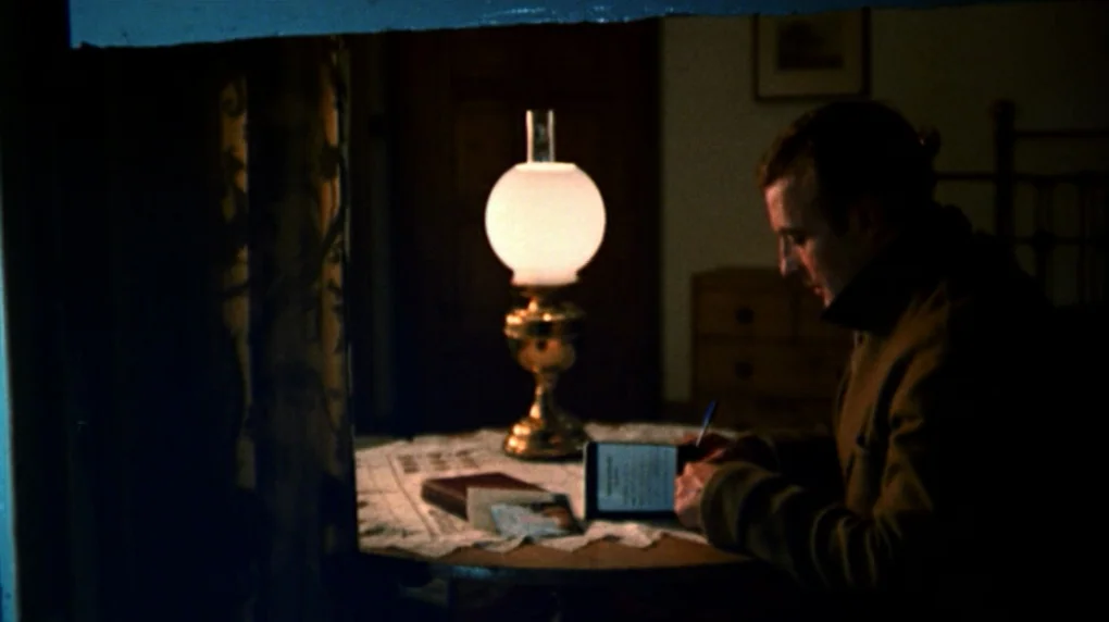 The Wicker Man movie still: medium — A dark, sparsely furnished room features a small table illuminated by a central lamp. A man sits at the table,…