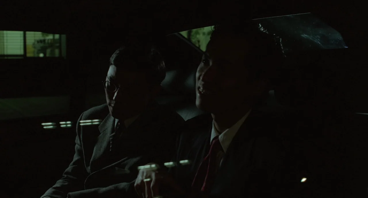 A Confucian Confusion (1994) movie still: medium — Two men in business attire sit in the dimly lit backseat of a moving vehicle. They are engaged in…
