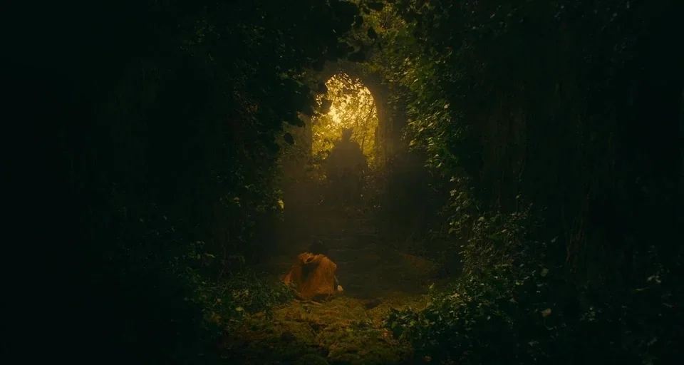The Green Knight (2021) movie still: wide — A dark stone archway overgrown with vines serves as a tunnel entrance. A person in a tan cloak sits on…