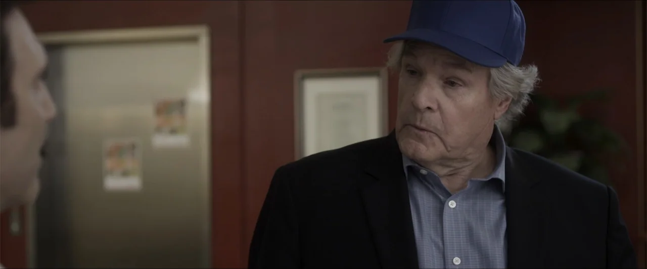 Heavenly Deposit (2019) movie still: medium — An interior office space with deep red walls and a metal door in view. An older man in a blue baseball…