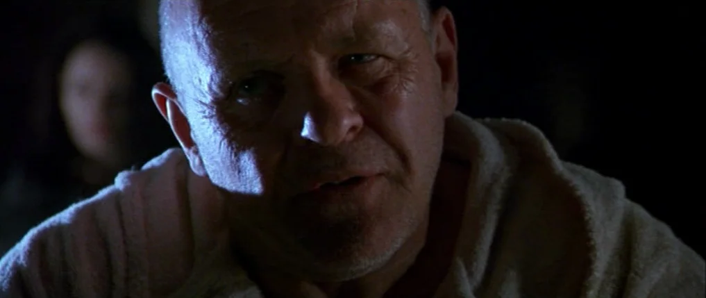 Titus (1999) movie still: closeup — A dark, dimly lit indoor space with a single light source highlighting a man. A bald man faces the camera…