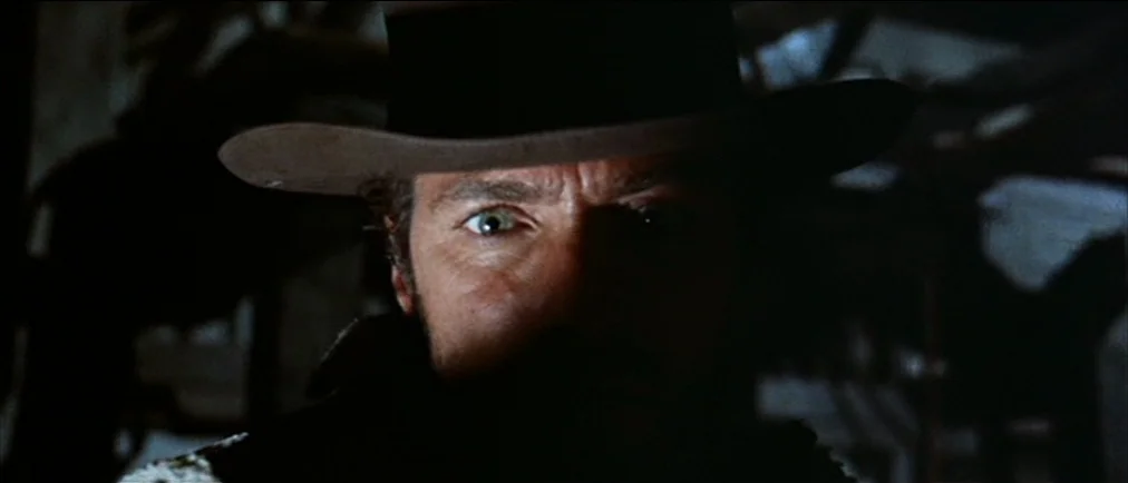 Fistful Of Dollars (1964) movie still: closeup — A dark wooden interior with obscure shapes in the background. A man stares forward while his face…