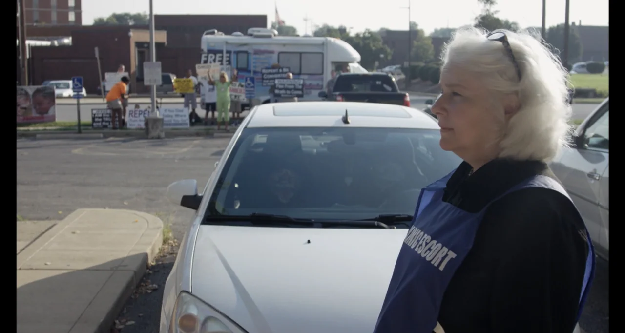 Abortion: Stories Women Tell (2016) movie still: medium — A paved lot features a white sedan parked with protestors visible in the background. An…