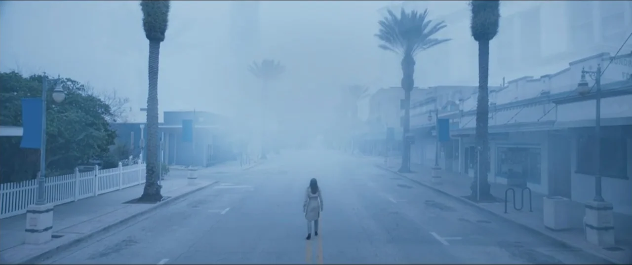 Offseason (2022) movie still: wide — An empty town street is filled with dense fog and lined with palm trees. A lone person in a light-colored coat…
