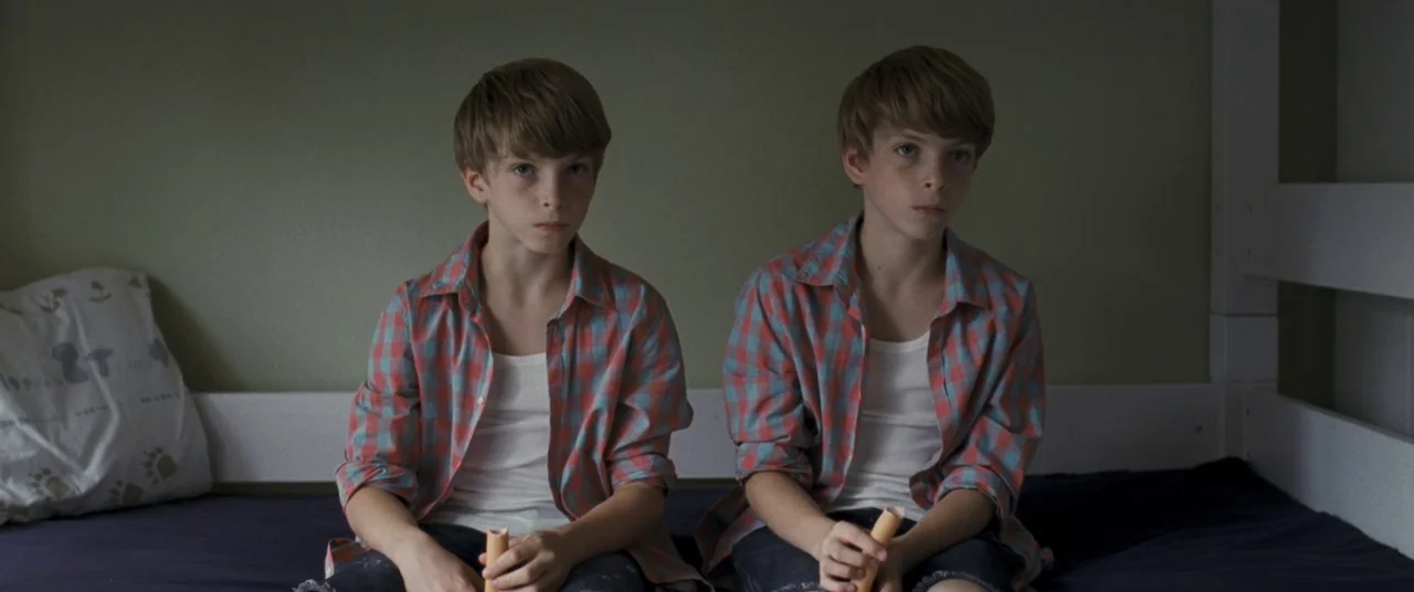 Goodnight Mommy (2014) movie still: medium — Two identical twin boys sit side by side on a mattress against a wall. Each boy wears an identical red…