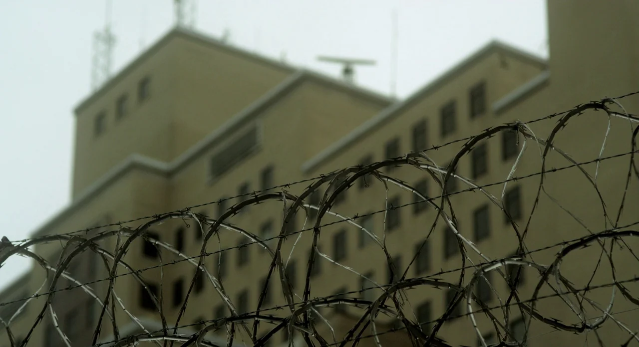 Side Effects (2013) movie still: establishing — A tall multi-story beige building sits under a flat, overcast sky. Layers of razor wire form a tight…
