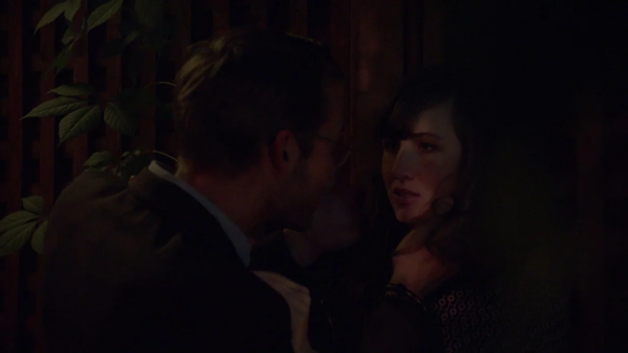 The Heart Machine (2014) movie still: medium — A dimly lit outdoor patio features a wooden trellis wall covered in climbing leaves. A man and a…