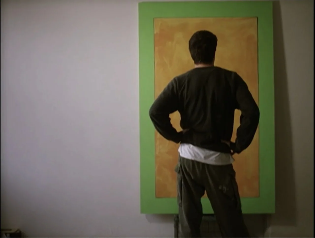 Apartment 12 (2001) movie still: medium — A plain room features a large rectangular painting with a bright green border. A man in casual clothes…