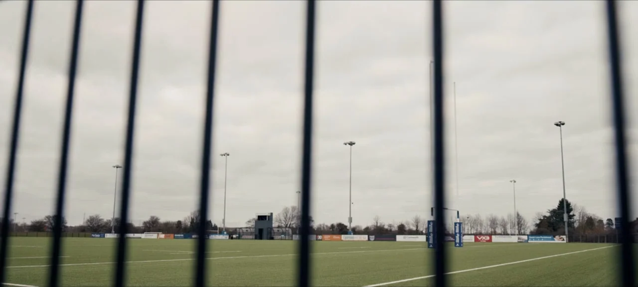 No Woman No Try (2022) movie still: wide — An empty synthetic turf pitch is framed behind vertical black metal security bars. Goalposts and sponsor…