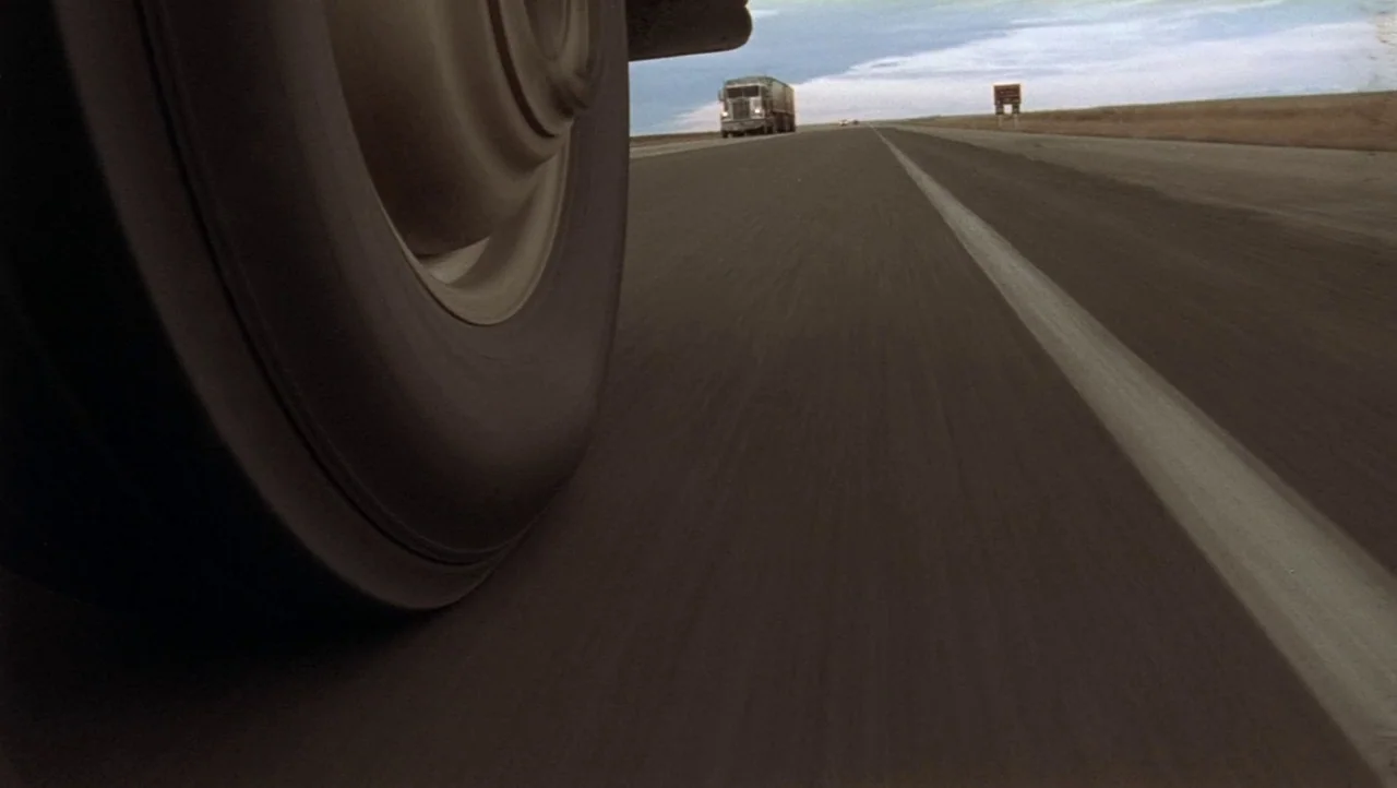 Hard Core Logo movie still: closeup — A low-angle view captures a large vehicle tire rolling along a flat highway. A truck is visible in the…