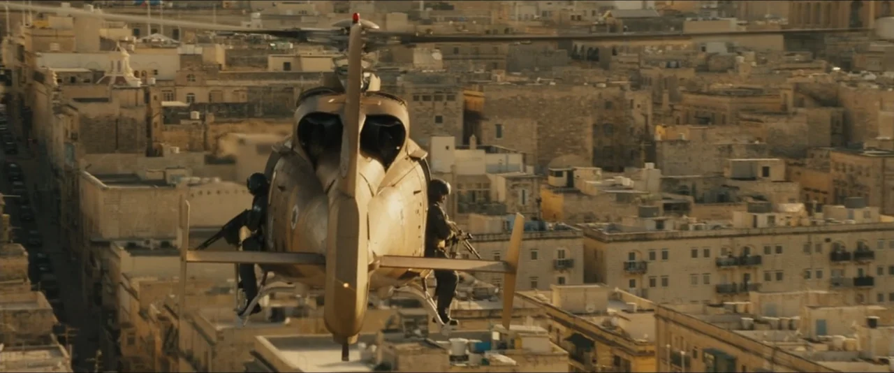 World War Z (2013) movie still: wide — A military helicopter flies at low altitude over a dense urban area with limestone buildings. Two armed…
